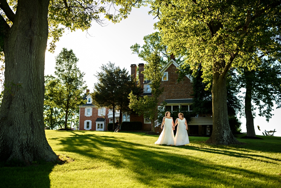 Two brides hold hands and walk across the grass at the eagle manor in NJ.
